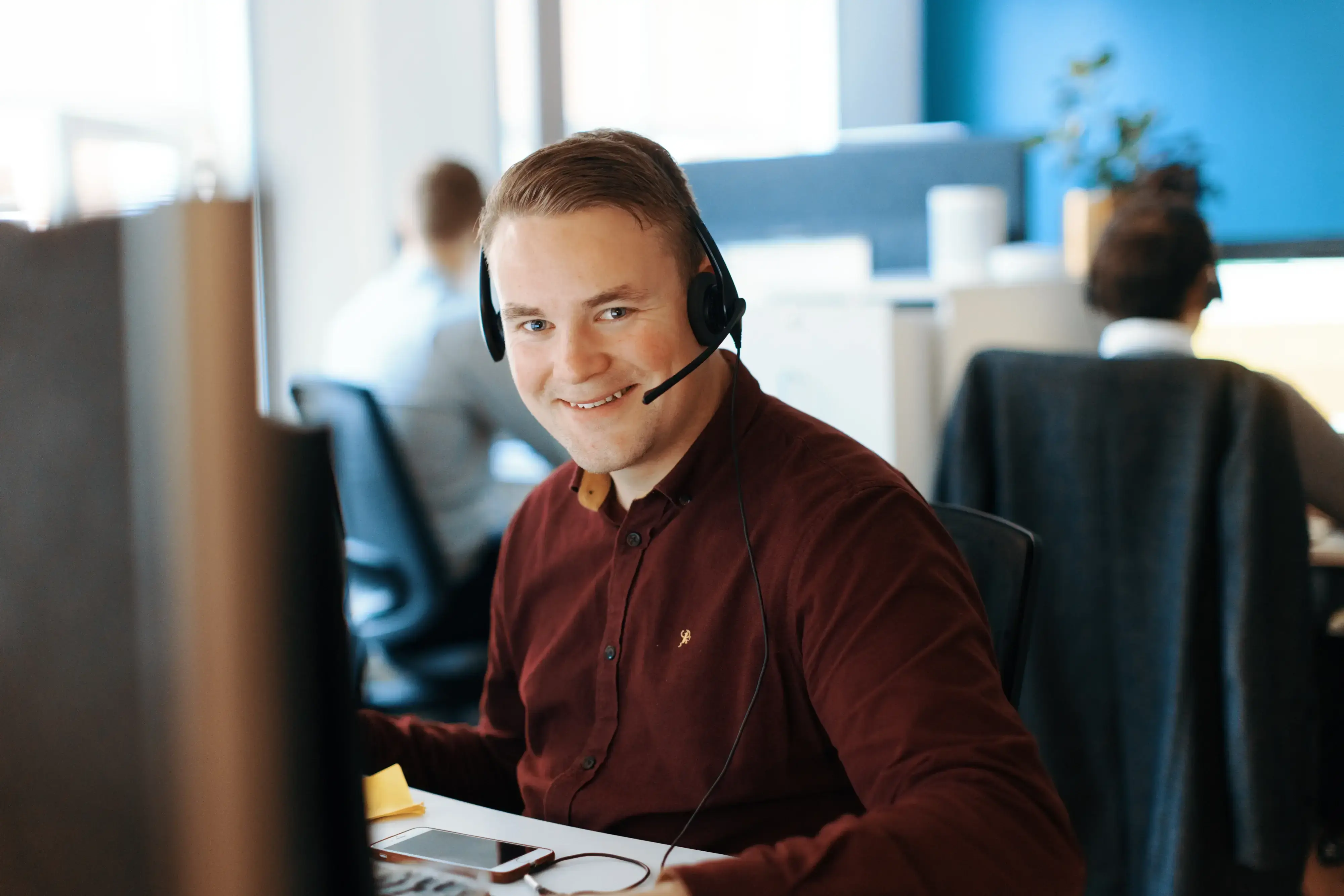 Smilende mann med headset sitter foran en dataskjerm i et åpent kontorlandskap, med kollegaer som jobber i bakgrunnen.