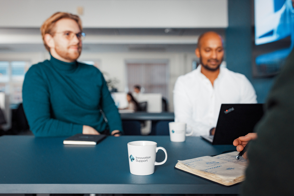To menn sitter ved et møtebord, den ene med en notatbok og den andre med en bærbar PC, i samtale med en person hvis hånd er synlig i forgrunnen. En kaffekopp med Innovation Support-logoen står på bordet